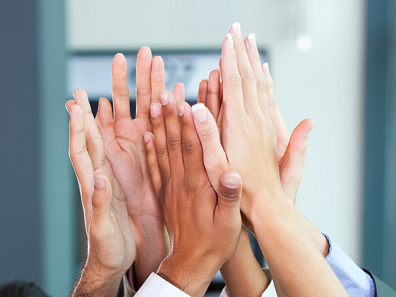 Auf dem Bild sind die Hände von sechs Personen zu sehen. Sie halten ihre Hände aneinander in der Luft.